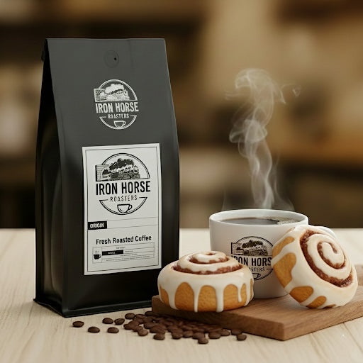 Black coffee bag with Iron Horse Roasters branding next to a steaming cup of coffee and cinnamon rolls on a wooden surface.